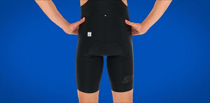 Back view of person wearing black Redux bib shorts against a blue background.