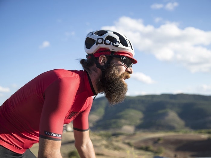 Bearded cyclist in a red jersey and white helmet riding outdoors with scenic hills in the background.