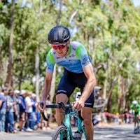 Cyclist in a blue and green jersey and black helmet racing through a tree-lined course with spectators in the background.