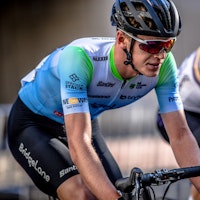 Cyclist in black bib shorts and a blue and green cycling jersey, riding on a wet road during a race, with other cyclists in the background.