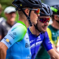 Cyclists at the starting line of a race, with a focus on a cyclist in a blue and green jersey and black helmet, alongside other riders in colorful jerseys.