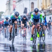 Cyclists in a blue and green cycling jersey and black bib shorts, racing on a wet street in rainy weather, with other cyclists in the background.