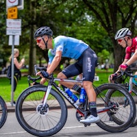 Cyclists riding in a park during a race, with one cyclist wearing a blue and green jersey and black bib shorts, and another cyclist in a red jersey.