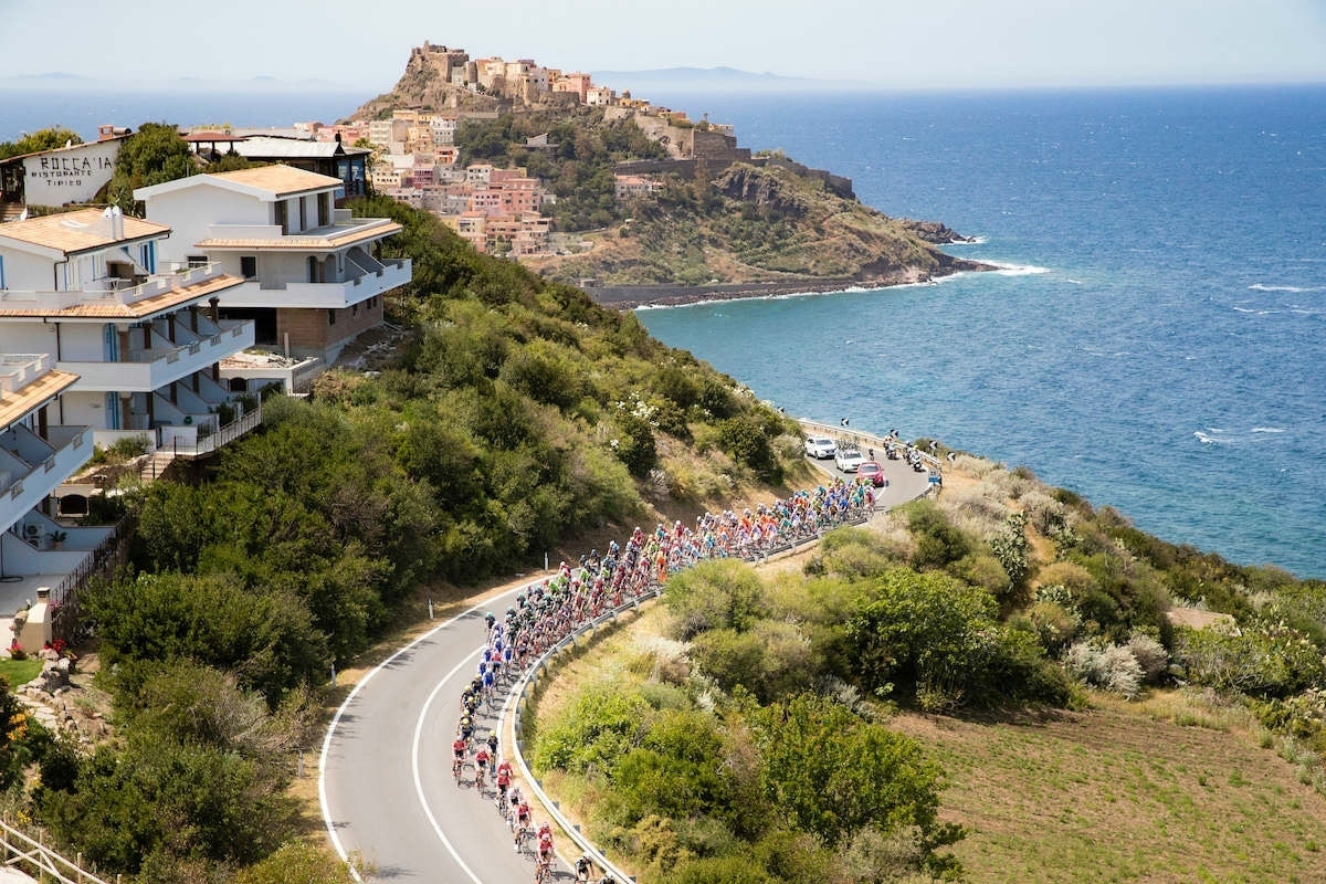 Cyclists riding on a coastal road with houses on the left and a scenic view of the ocean and a distant town on a hilltop.