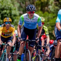 Cyclists competing in a race, with the focus on a cyclist in a blue and green jersey and black bib shorts, surrounded by other riders.