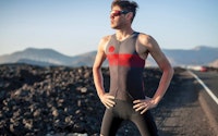 Triathlete standing on a road with mountains in the background, wearing a red and black triathlon suit.